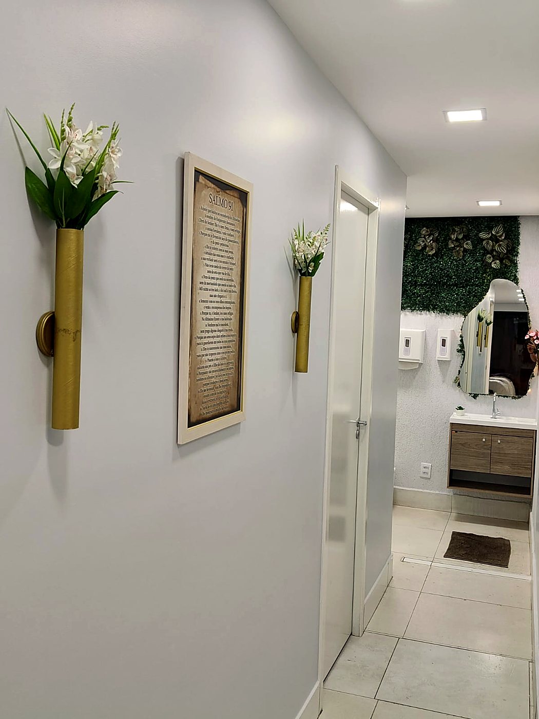 Corredor com vasos dourados e flores, com o lavabo de parede verde ao fundo