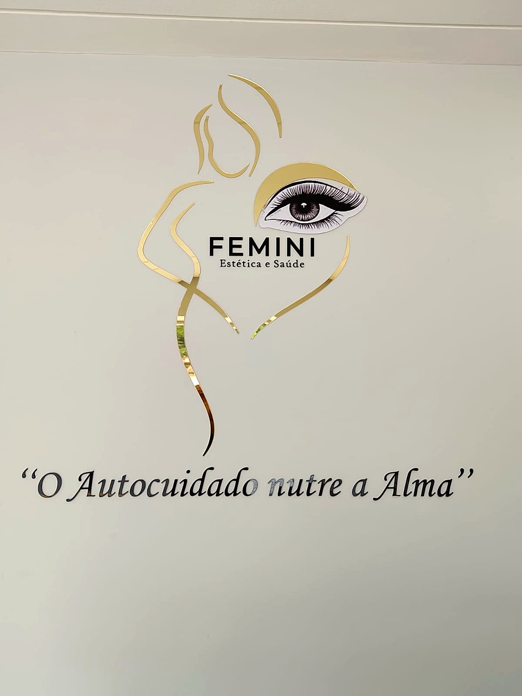 Logo FEMINI em acrílico dourado na parede, com a frase O Autocuidado nutre a Alma